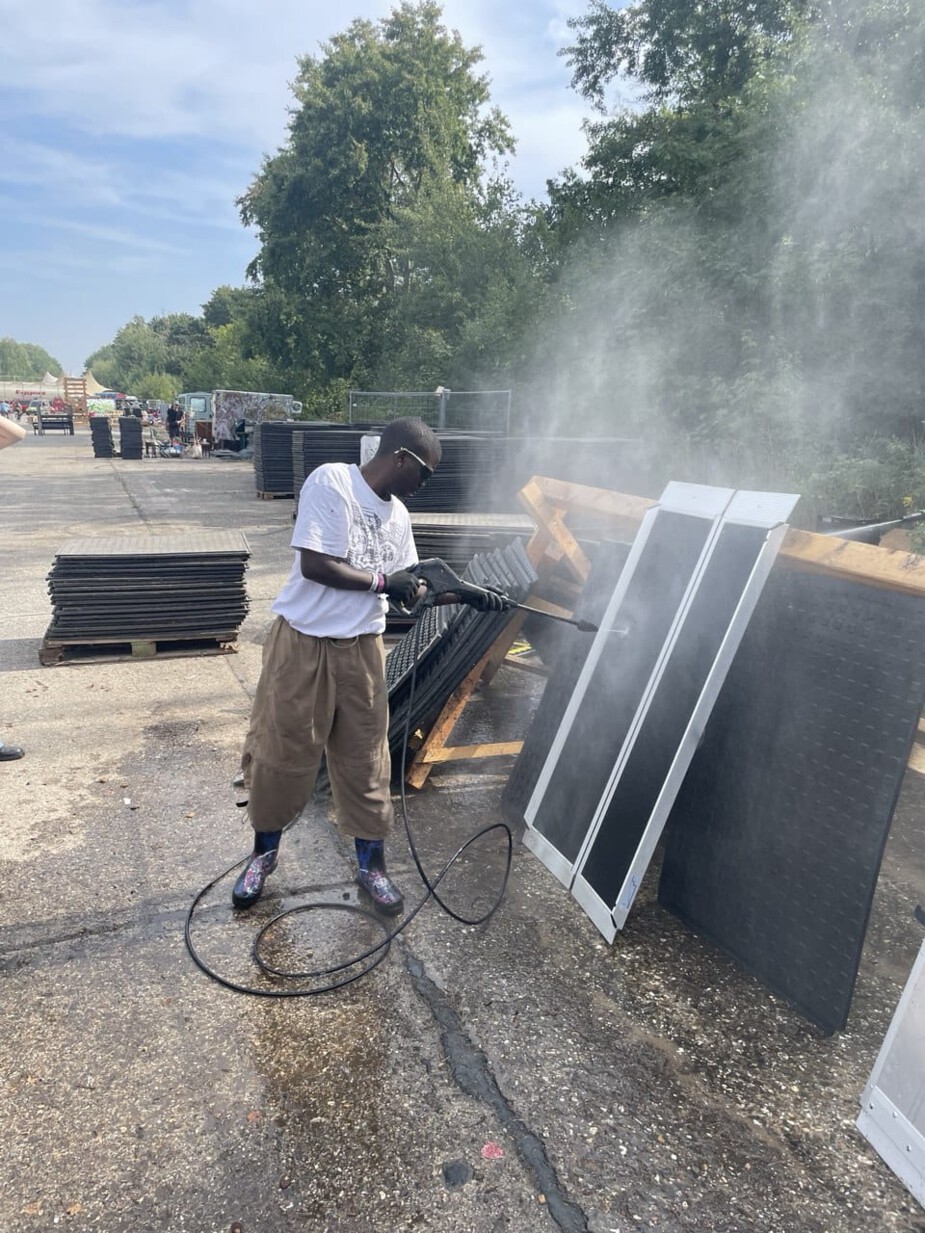 The picture shows the pressure wash station. A person is cleaning a metal ramp with a pressure washer. Singular heavy-duty mats are leaning against the left and right sides of the washing station.