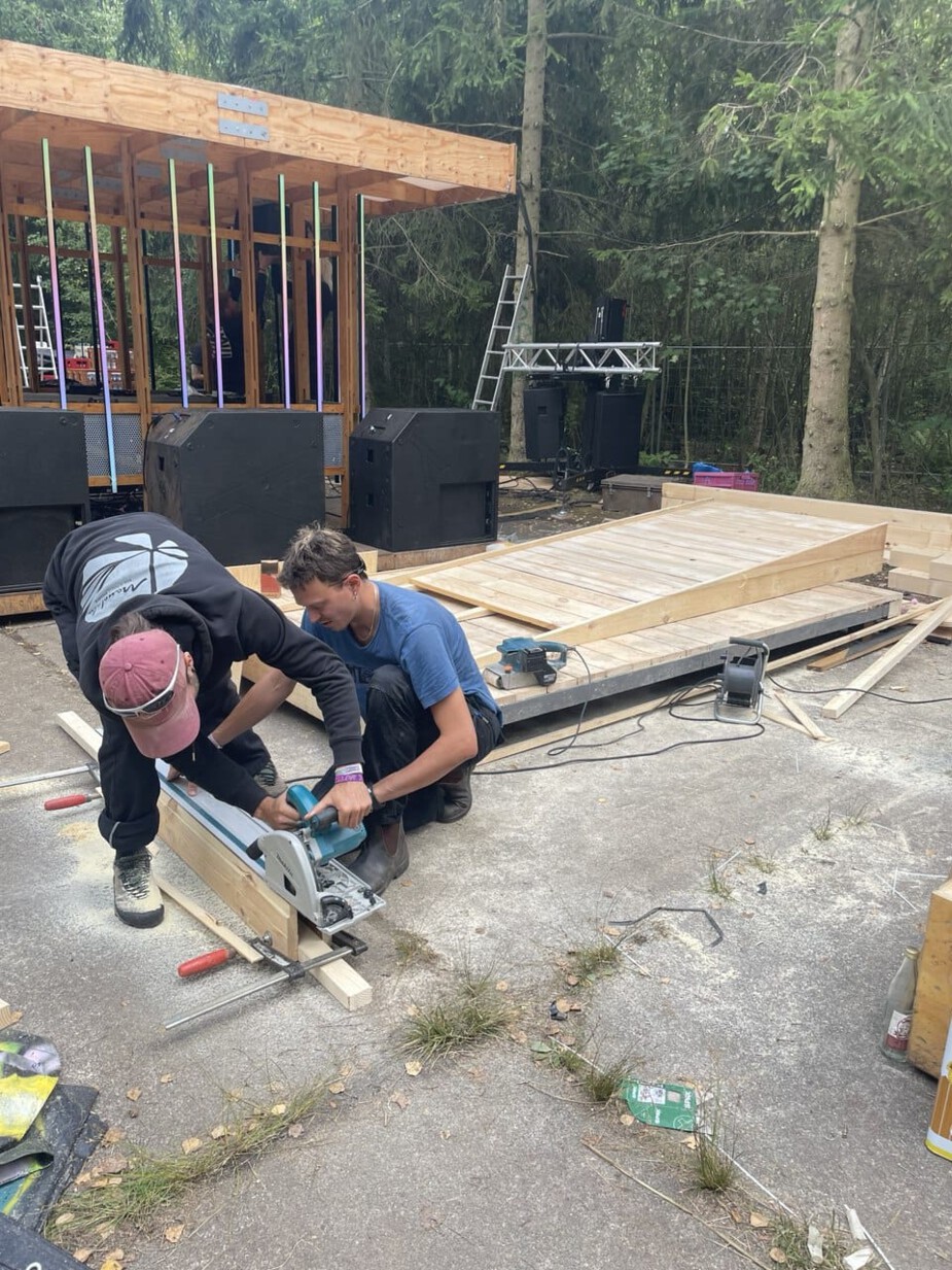 2 Männer hantieren mit Holz. im Hintergrund ist ein Podest und eine Rampe aus Holz.